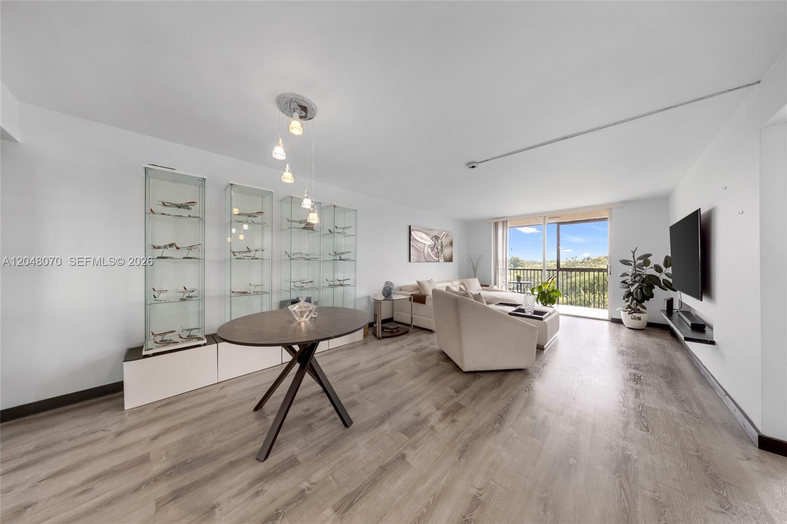 A living room features light wood-look flooring, glass display cabinets, a dining table, and balcony access — 3001 W Rolling Hills Cir # 610, Davie FL 33328, DAVIE, FL
