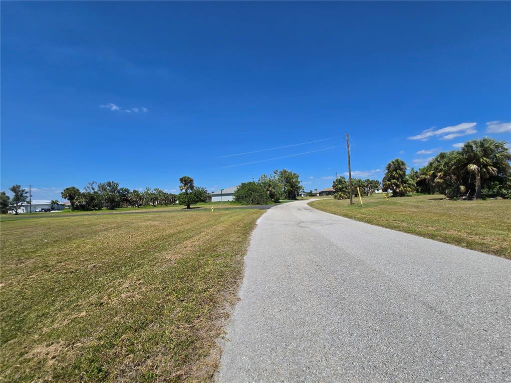 A paved road winds through a grassy, open lot with scattered trees under a blue sky — 16284 JUAREZ COURT, PUNTA GORDA, FL