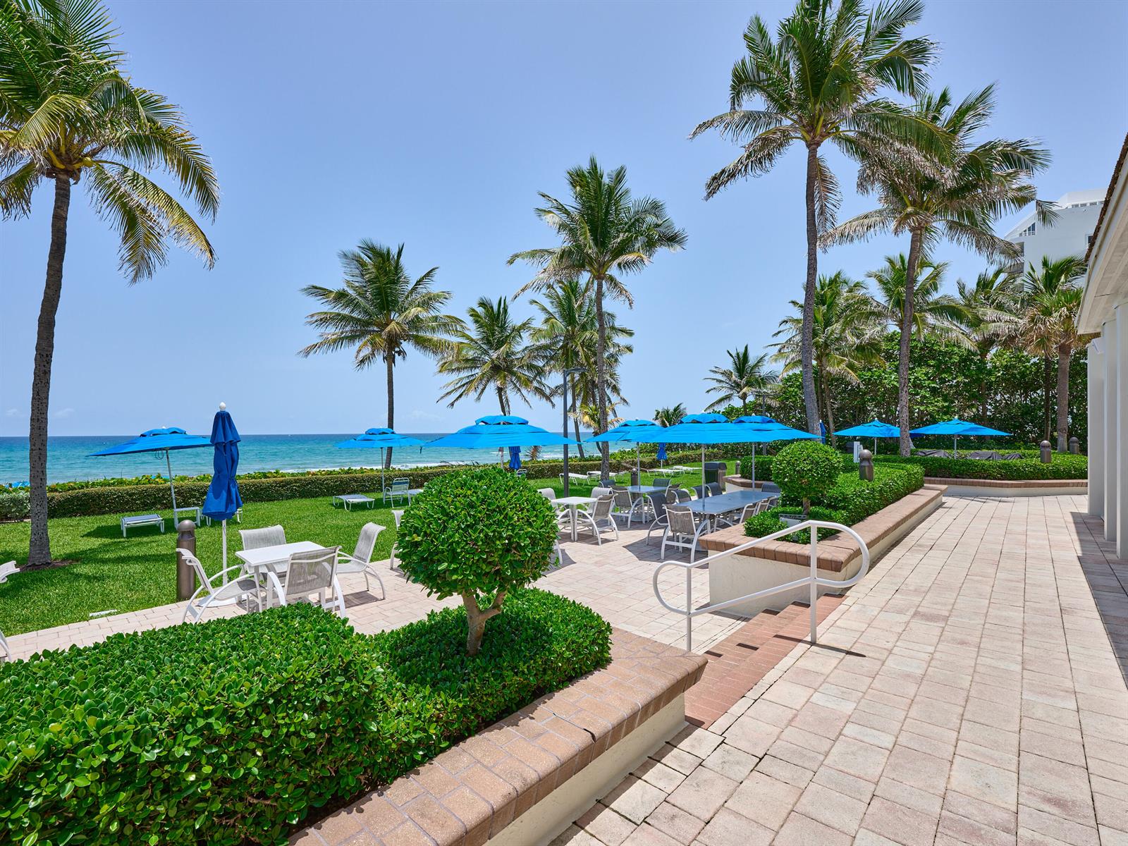 An outdoor patio with tables, chairs, and blue umbrellas overlooks a grassy lawn and ocean — 3420 S Ocean Boulevard, Highland Beach FL 33487, HIGHLAND BEACH, FL