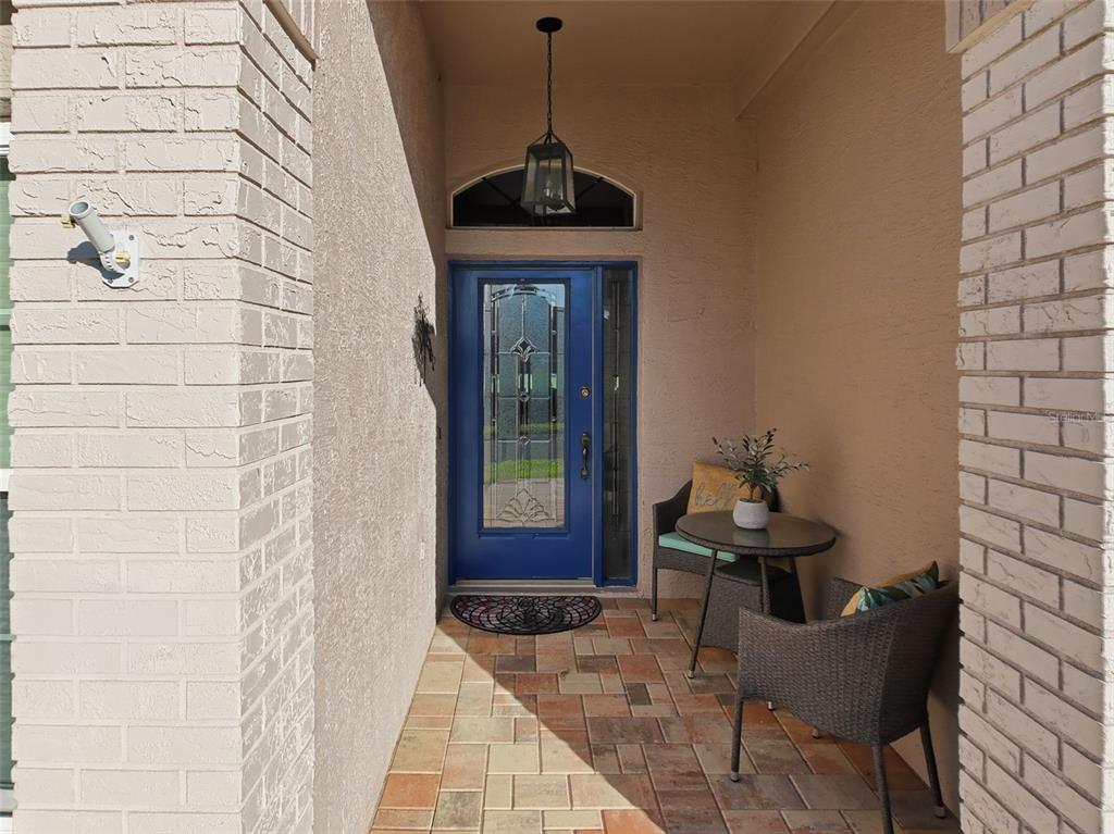 A covered front porch features a blue door, brick columns, and a small bistro set — 7442 WOODHOLLOW ROAD, SPRING HILL, FL
