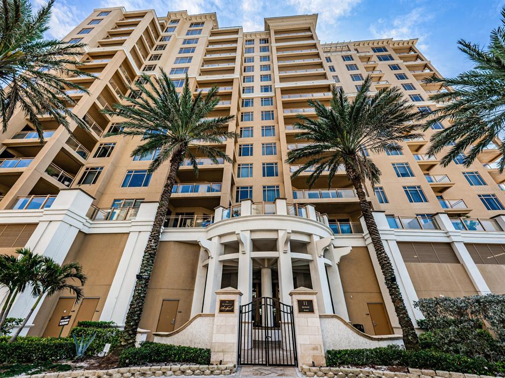 A low-angle view of a multi-story tan apartment building with palm trees and gated entrance — 11 BAYMONT STREET UNIT 403, CLEARWATER BEACH, FL
