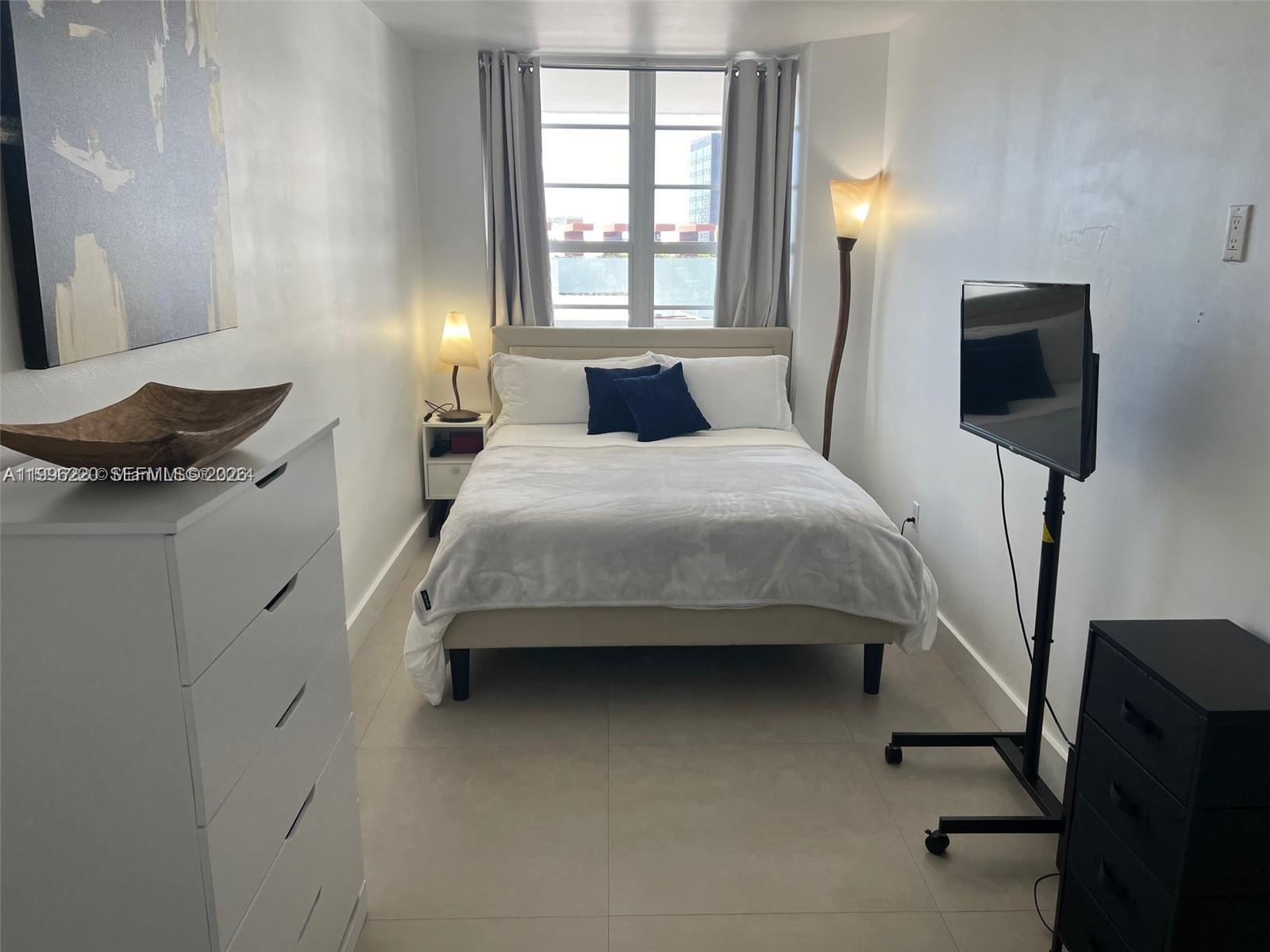 A bedroom features a bed, white dresser, rolling television stand, and a window with curtains — 100 Lincoln Rd # 620, Miami Beach FL 33139, MIAMI BEACH, FL