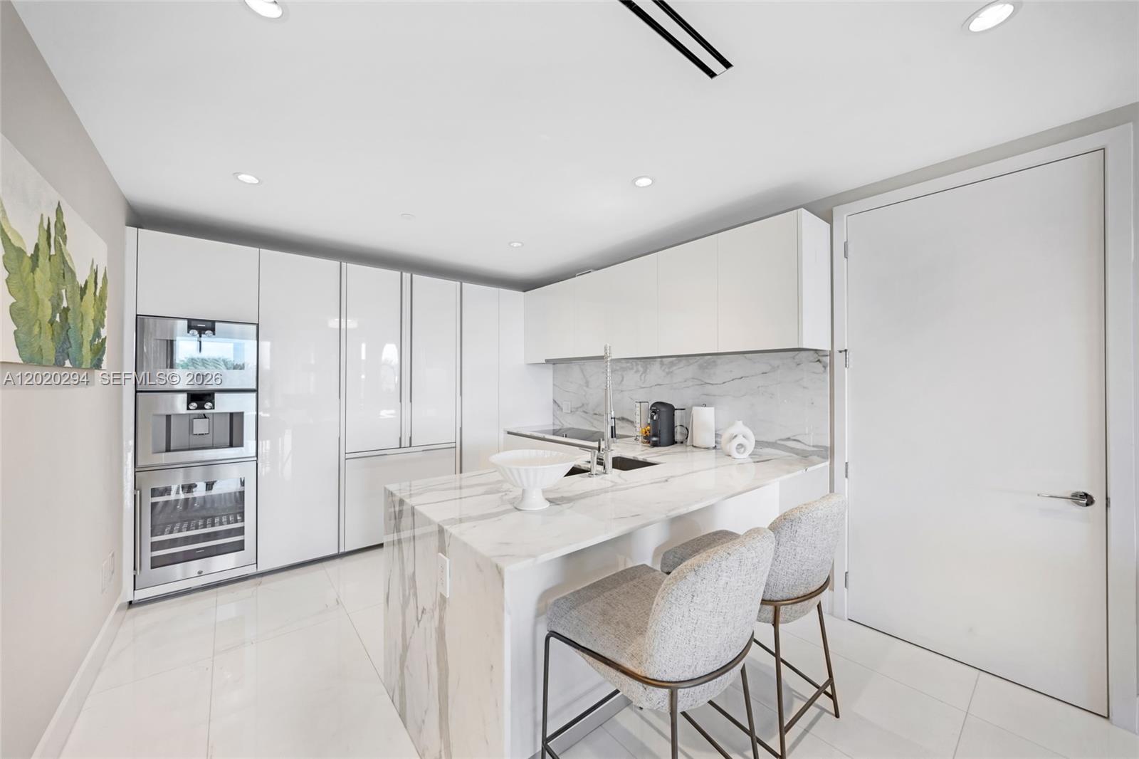 A modern kitchen features white cabinetry, marble countertops, and a waterfall island with bar stools — 10201 Collins Ave # 305, Bal Harbour FL 33154, BAL HARBOUR, FL