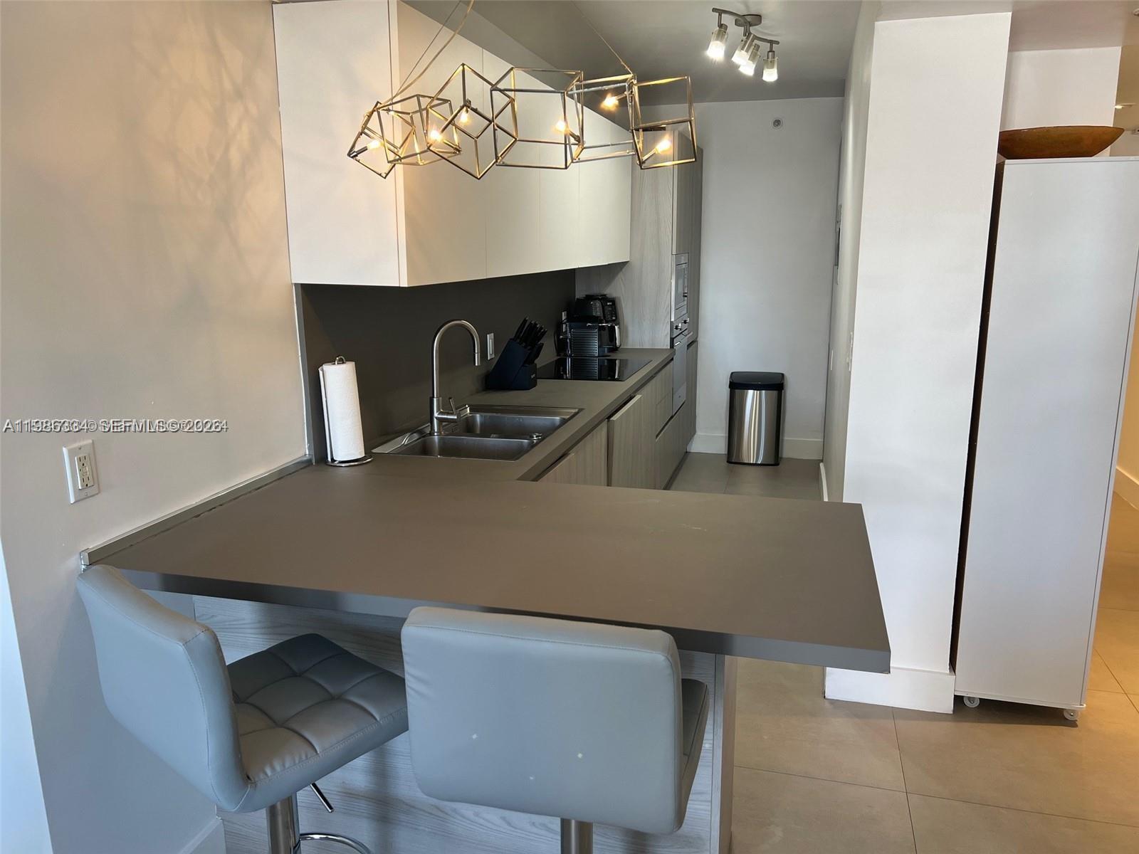 A modern kitchen features gray countertops, white cabinetry, and a breakfast bar with two stools — 100 Lincoln Rd # 620, Miami Beach FL 33139, MIAMI BEACH, FL