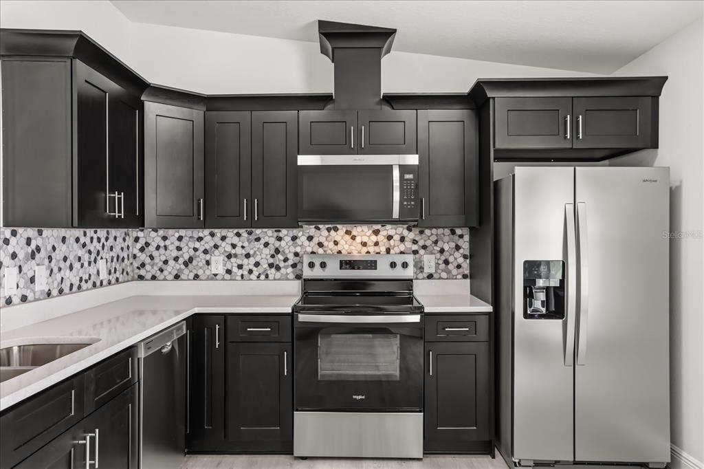 A kitchen features dark cabinets, stainless steel appliances, white countertops, and a pebble-patterned backsplash — 710 BEECHER STREET, LEESBURG, FL