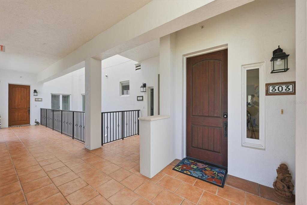 An outdoor hallway with tiled floors leads to a wooden front door labeled 303 — 11160 HACIENDA DEL MAR BOULEVARD UNIT D-303, PLACIDA, FL
