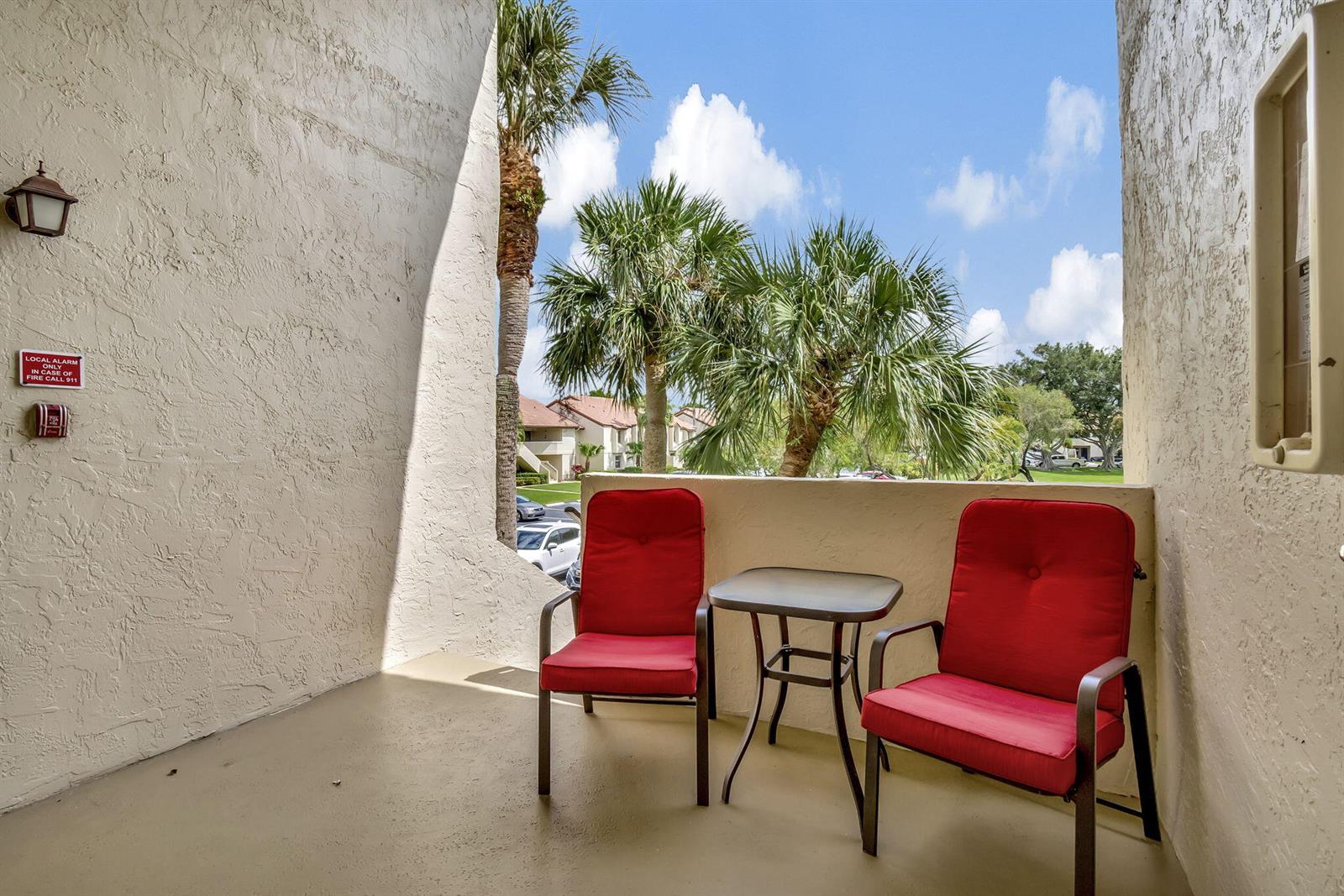 A balcony features two red chairs and a small table overlooking palm trees and buildings — 5869 Parkwalk Drive, Boynton Beach FL 33472, BOYNTON BEACH, FL