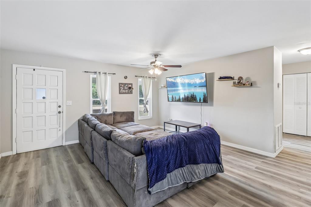 A living room features gray wood-look flooring, a large sectional sofa, and a wall-mounted television — 1200 E HANCOCK DRIVE, DELTONA, FL