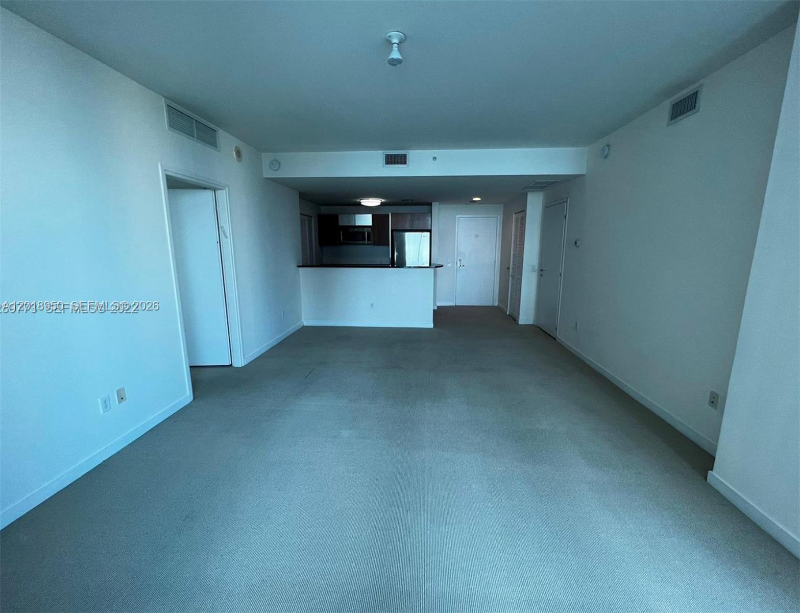 An empty living area with beige carpeting leads to an open kitchen with dark cabinetry — 300 S Biscayne Blvd # 1606, Miami FL 33131, MIAMI, FL