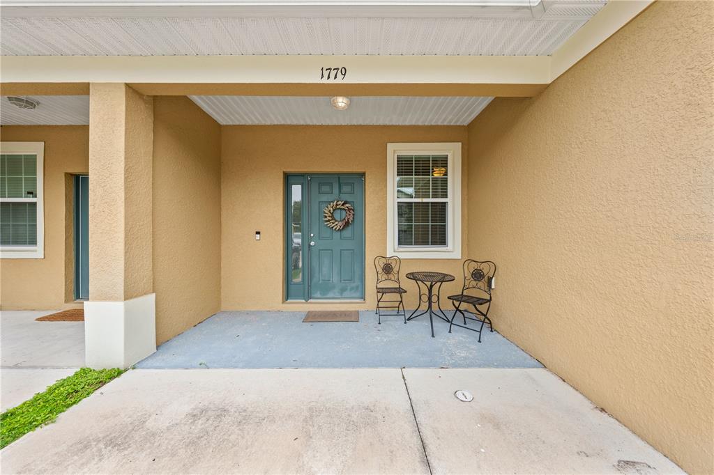 A covered concrete porch features a teal front door, a small bistro set, and stucco walls — 1779 BARTON TOWNE CIRCLE, ST CLOUD, FL