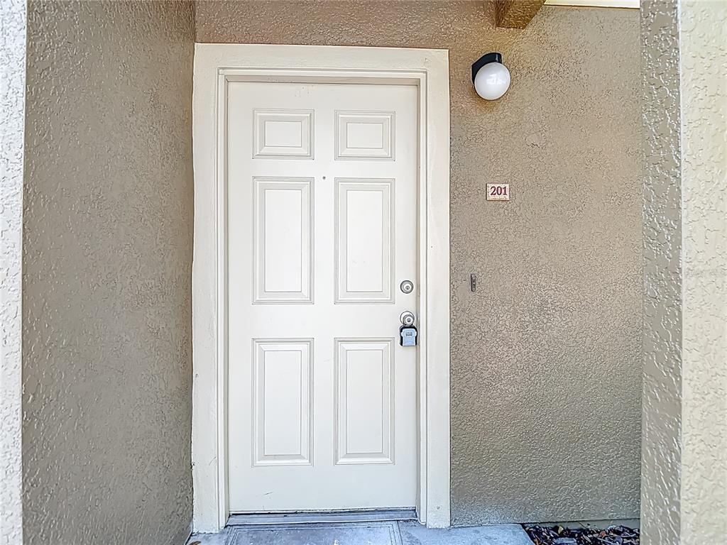 A white six-panel front door with a lockbox is set in a beige stucco wall — 36106 LAKE CHASE BOULEVARD Unit 201, ZEPHYRHILLS, FL
