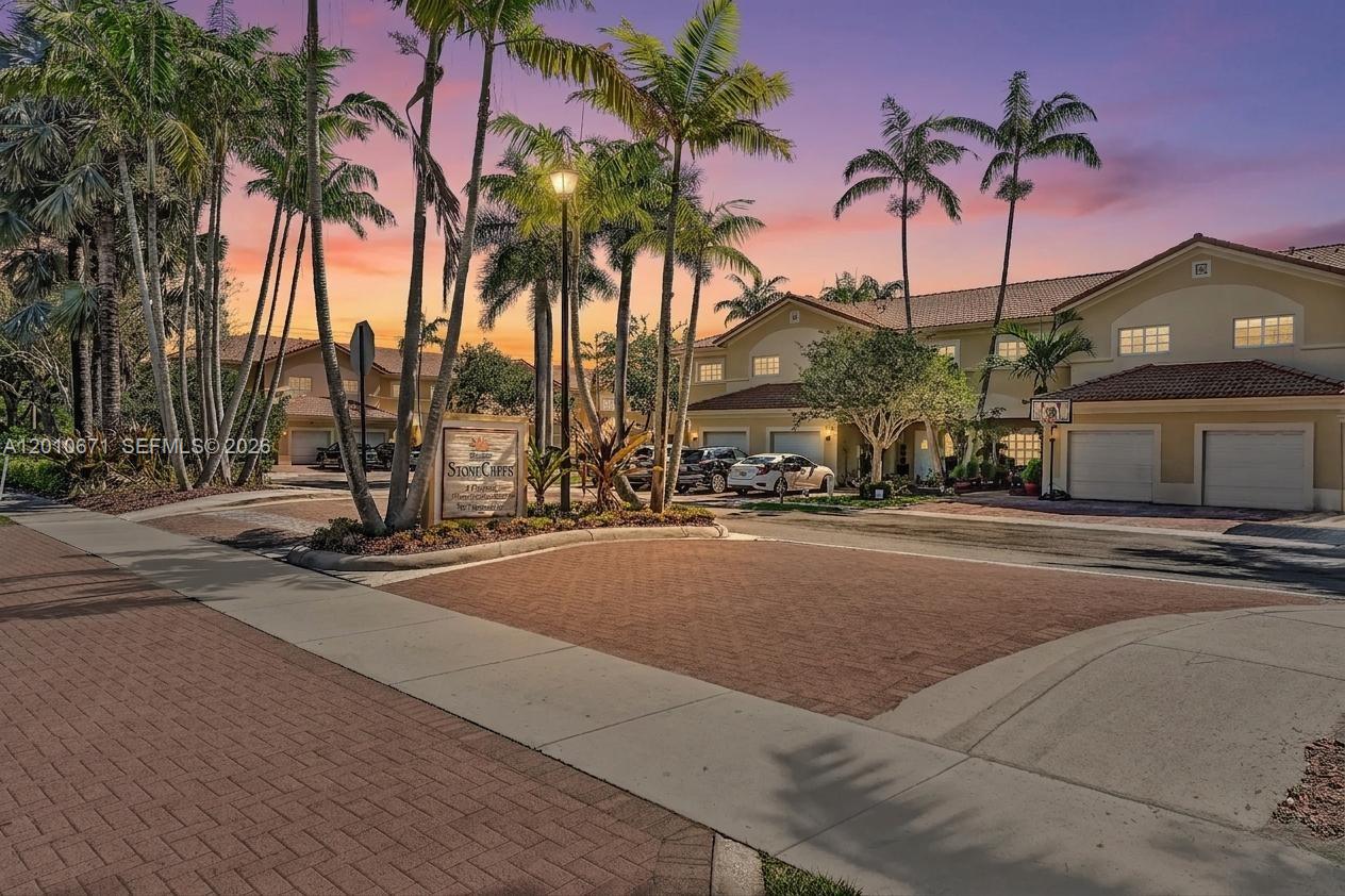 A residential community entrance features a stone sign, palm trees, and multi-story townhomes at sunset — 7659 N Stonecreek Cir, Davie FL 33024, DAVIE, FL