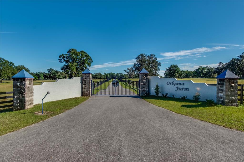 A paved driveway leads to a gated entrance with stone pillars and white walls — 7787 NW 150TH AVENUE, MORRISTON, FL