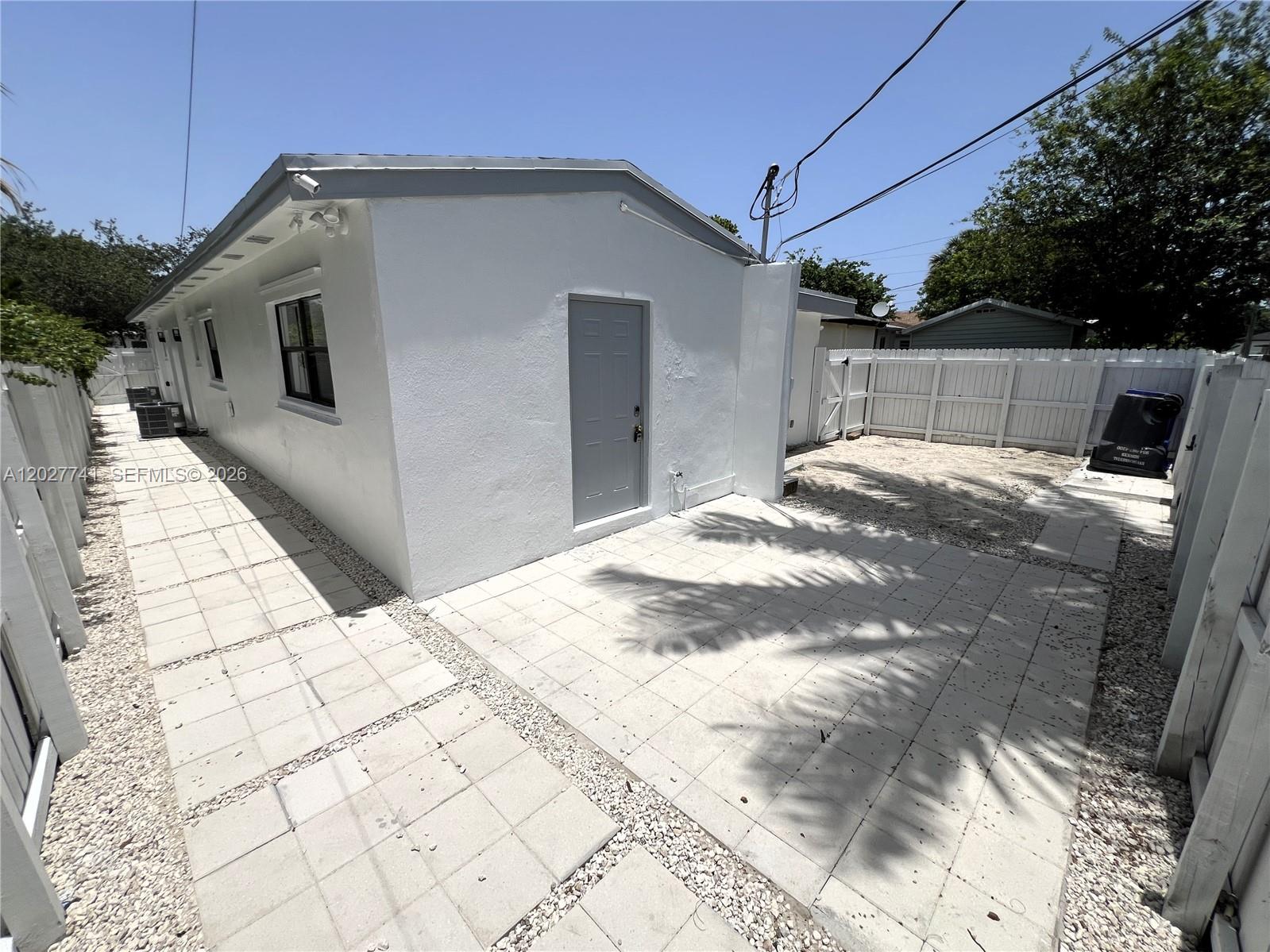 A white exterior wall features a gray door, a paved patio, and a white fence — 2226 Cody St, Hollywood FL 33020, HOLLYWOOD, FL