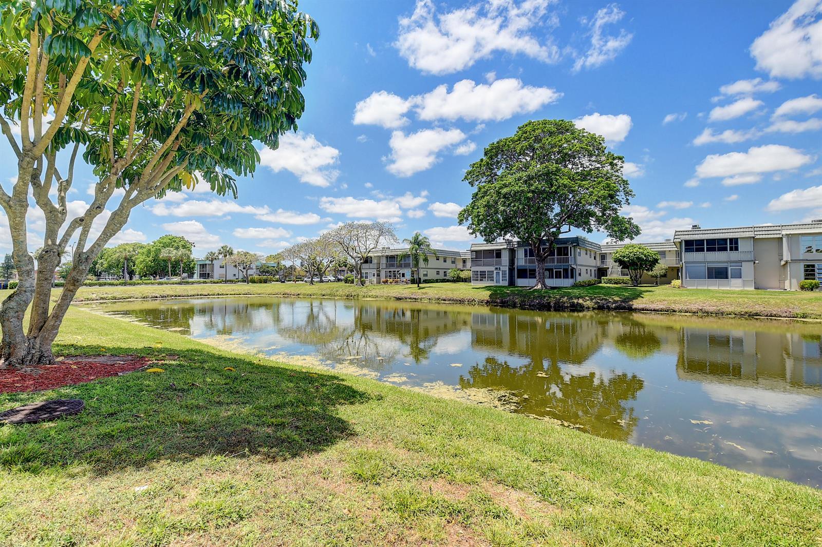 A pond sits in front of apartment buildings under a blue sky with scattered clouds — 318 Monaco G # G, Delray Beach FL 33446, DELRAY BEACH, FL