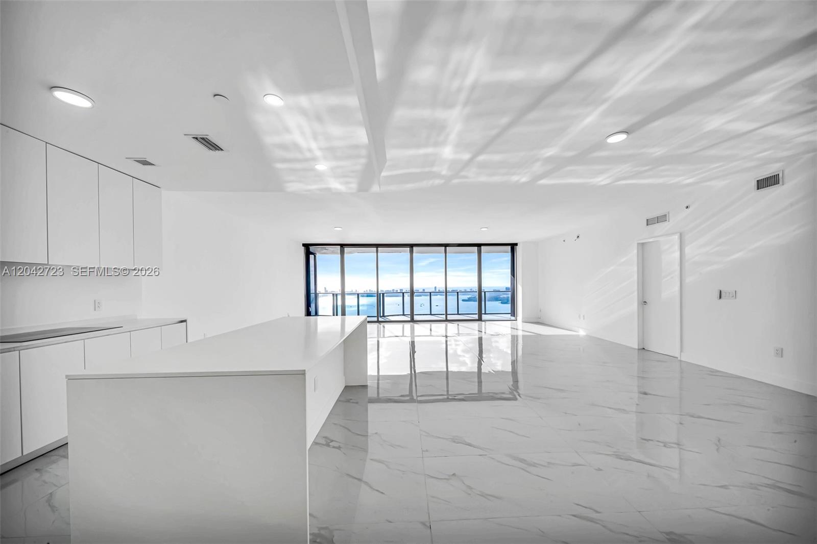 A modern, minimalist kitchen and living area features white cabinetry, a large island, and marble flooring — 700 NE 24th St # 4702, Miami FL 33137, MIAMI, FL