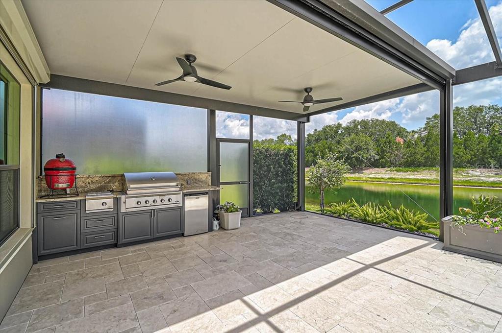 A covered outdoor kitchen with a grill, kamado smoker, and stone flooring overlooks a pond — 14911 CASTLE PARK TERRACE, LAKEWOOD RANCH, FL