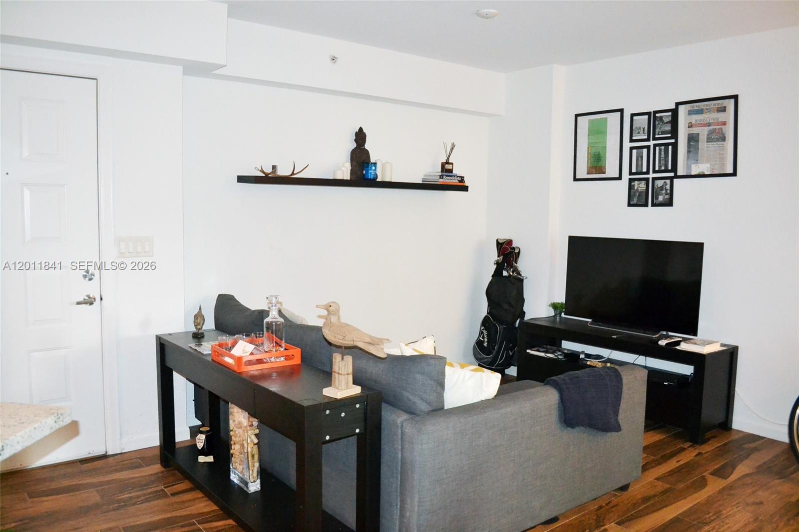 A living room features a grey sofa, a console table, a television, and wall-mounted shelving — 1830 Radius Dr # 317, Hollywood FL 33020, HOLLYWOOD, FL