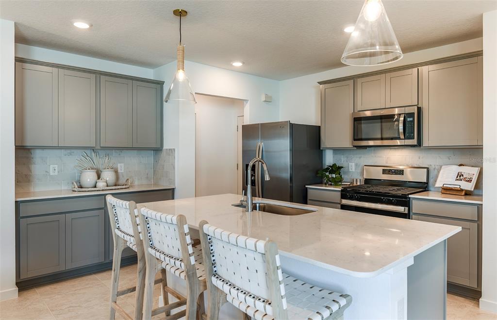 A modern kitchen features gray cabinets, a white quartz island with seating, and stainless appliances — 1391 ALLAGASH DRIVE, ZEPHYRHILLS, FL