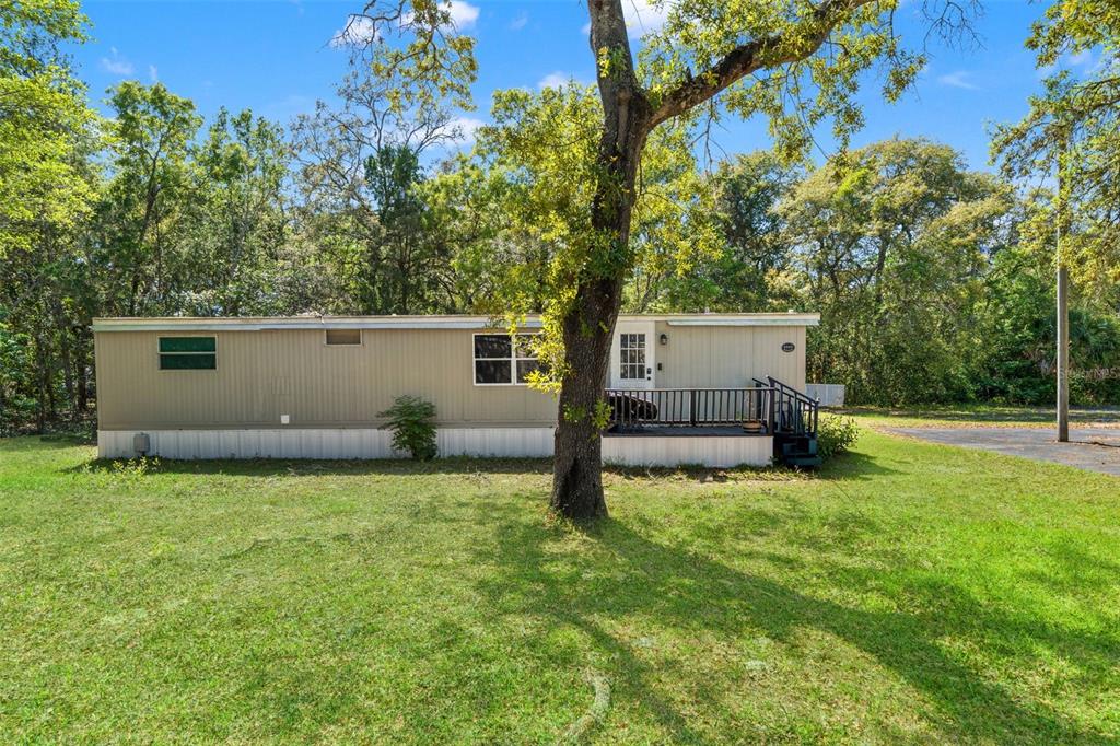 A single-story mobile home with a small front deck sits on a grassy, wooded lot — 3345 BALLARD STREET, SPRING HILL, FL