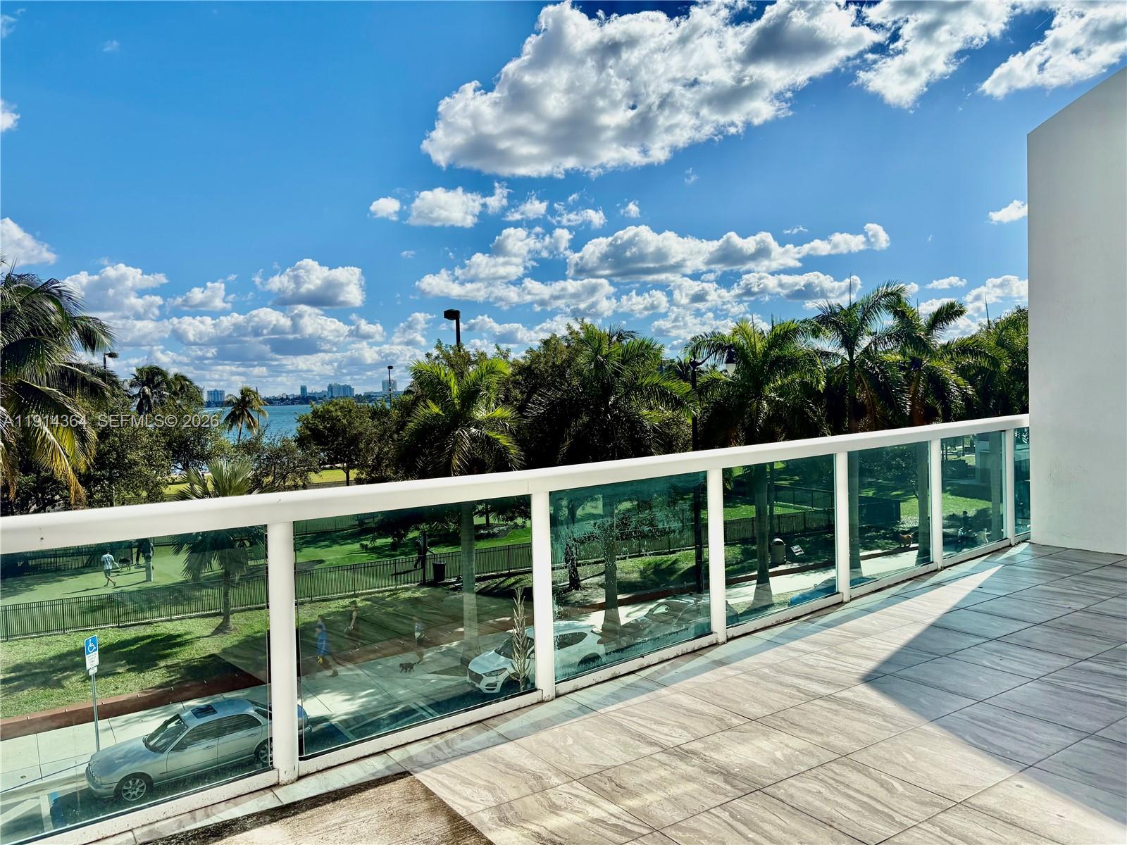 A balcony with glass railings overlooks a park, palm trees, and a distant waterfront — 1800 N Bayshore Dr # 306, Miami FL 33132, MIAMI, FL