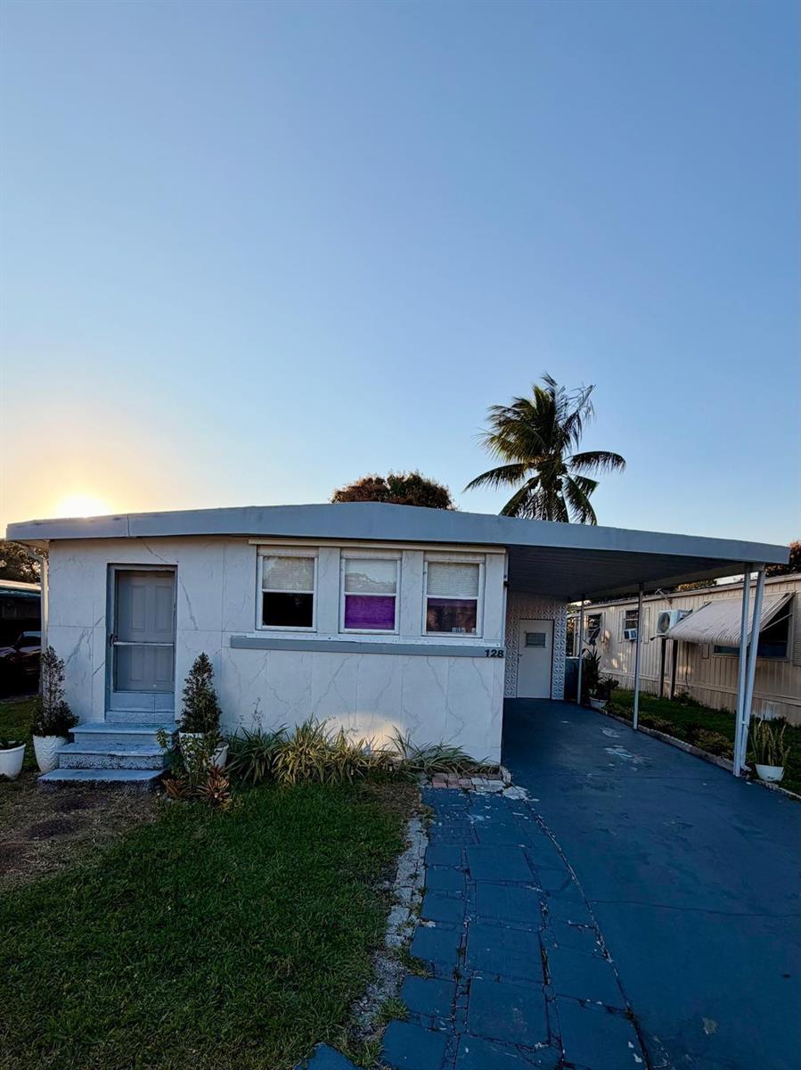 A single-story mobile home with a carport and a small front yard at sunset — 128 Havenwood Drive, Pompano Beach FL 33064, POMPANO BEACH, FL