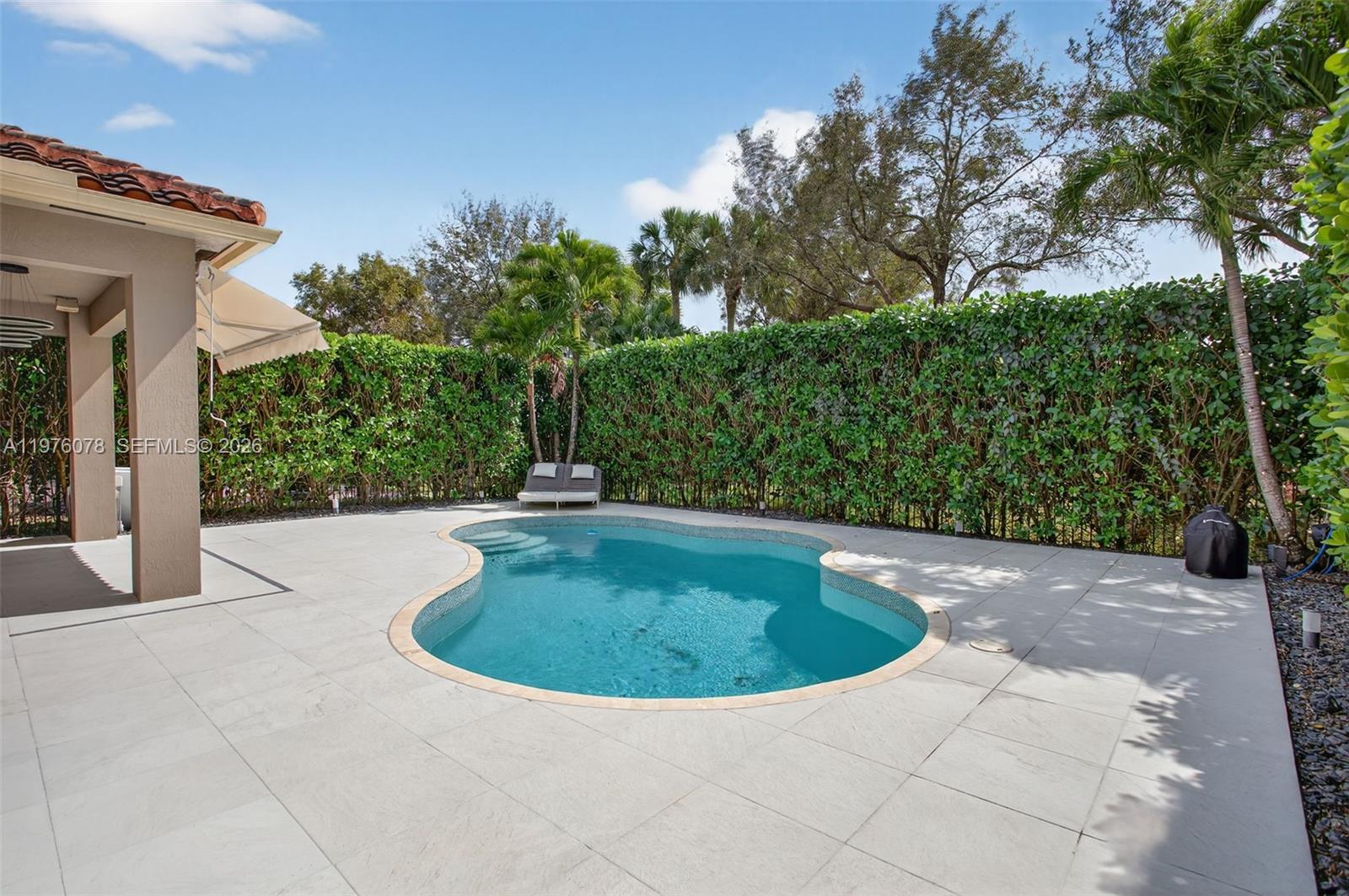 A kidney-shaped swimming pool sits on a paved patio surrounded by a tall privacy hedge — 11174 NW 72nd Pl, Parkland FL 33076, PARKLAND, FL