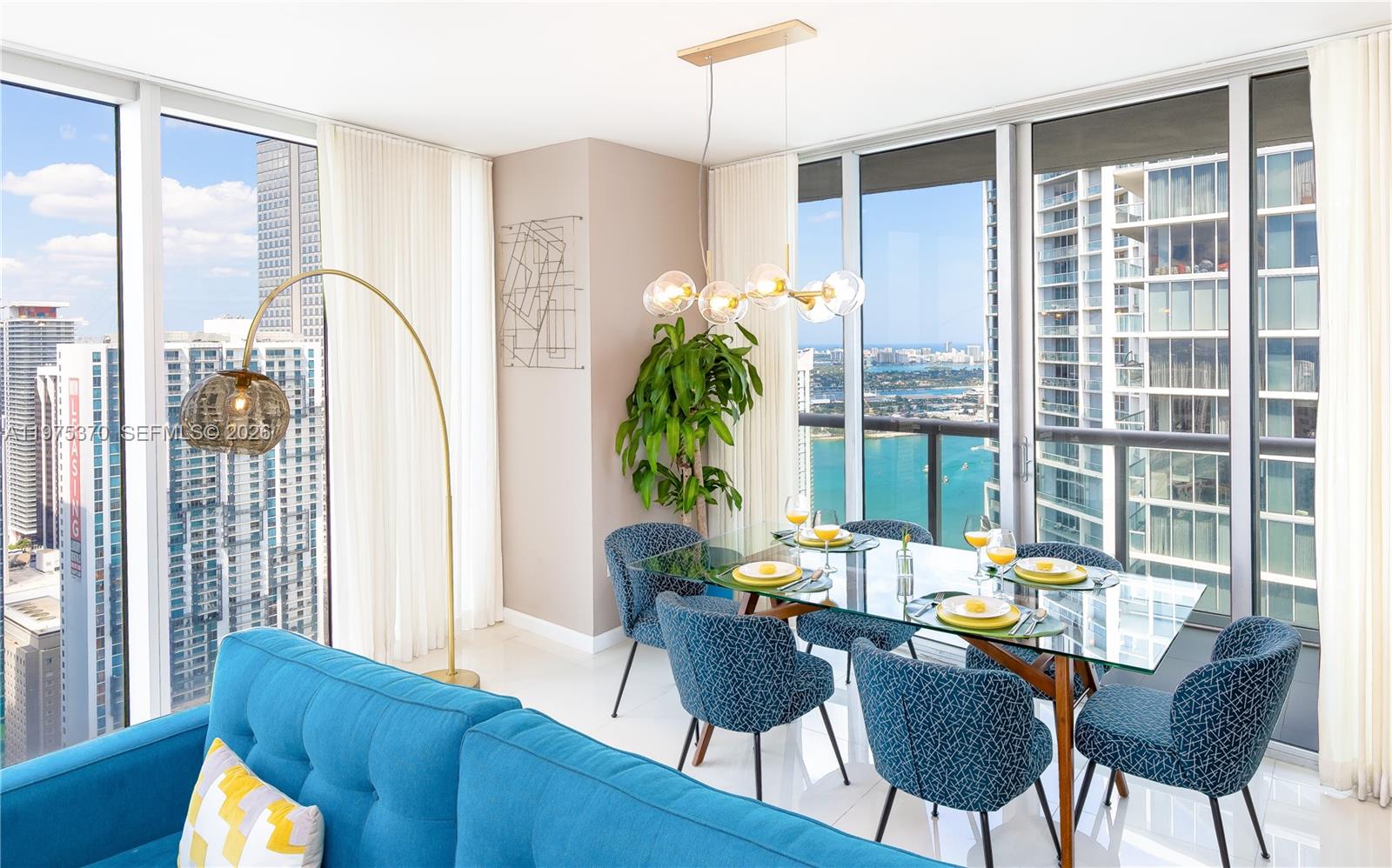 A dining area with a glass table and blue chairs overlooks a coastal city skyline — 485 Brickell Ave # 4702, Miami FL 33131, MIAMI, FL
