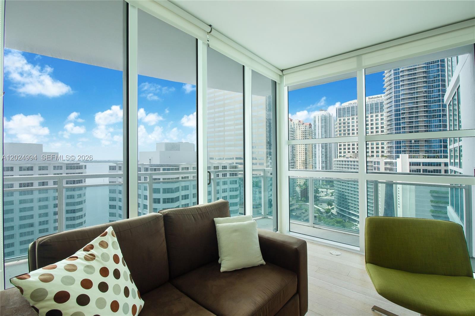 A living room features a brown sofa, a green chair, and floor-to-ceiling windows overlooking city buildings — 950 Brickell Bay Dr # 1911, Miami FL 33131, MIAMI, FL