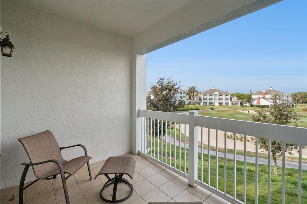 A covered balcony features tile flooring, a chair, an ottoman, and a golf course view — 7655 HERITAGE CROSSING WAY UNIT 201, REUNION, FL