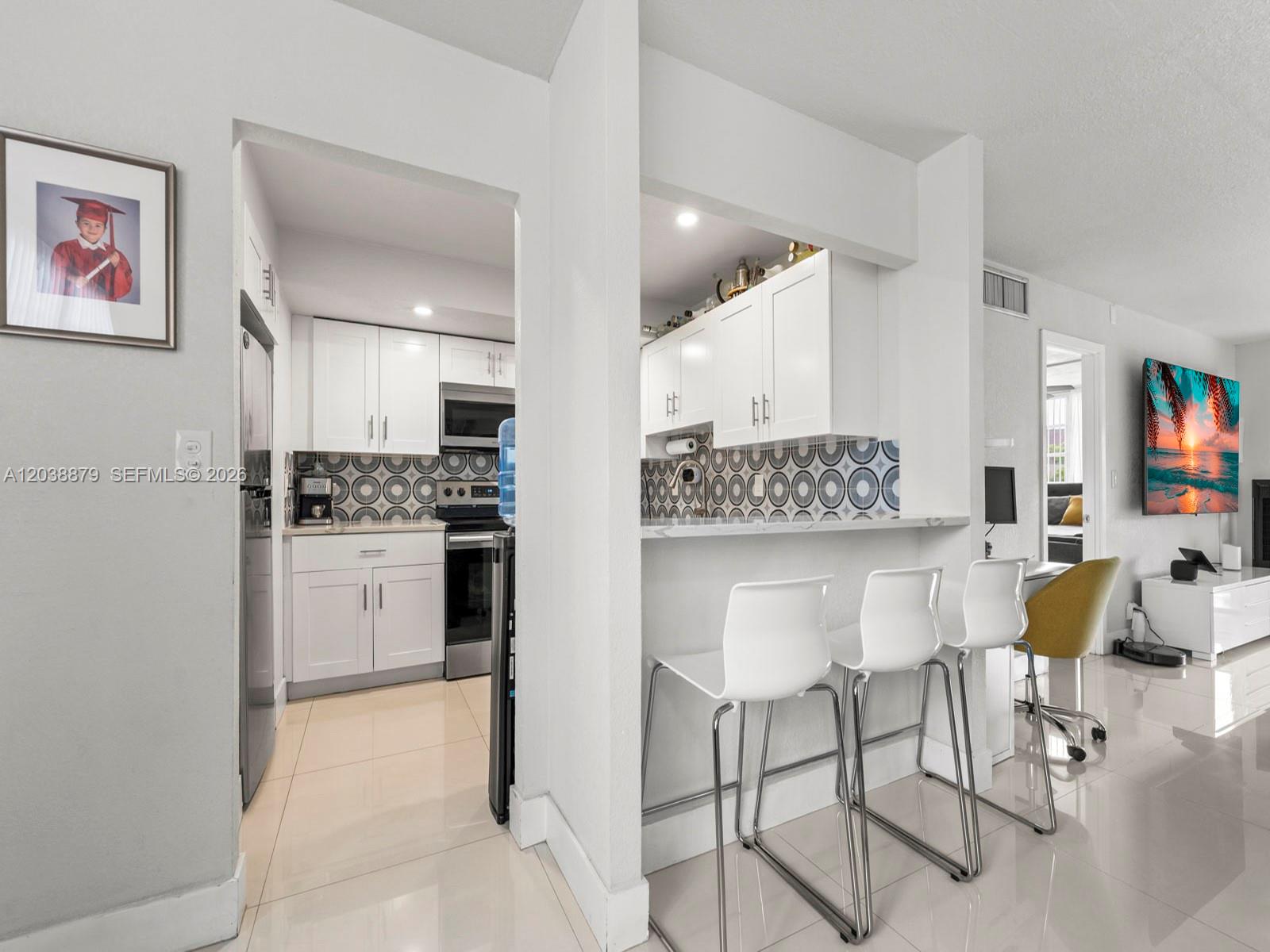 A kitchen with white cabinets and patterned backsplash opens to a breakfast bar with stools — 7021 Environ Blvd # 320, Lauderhill FL 33319, LAUDERHILL, FL
