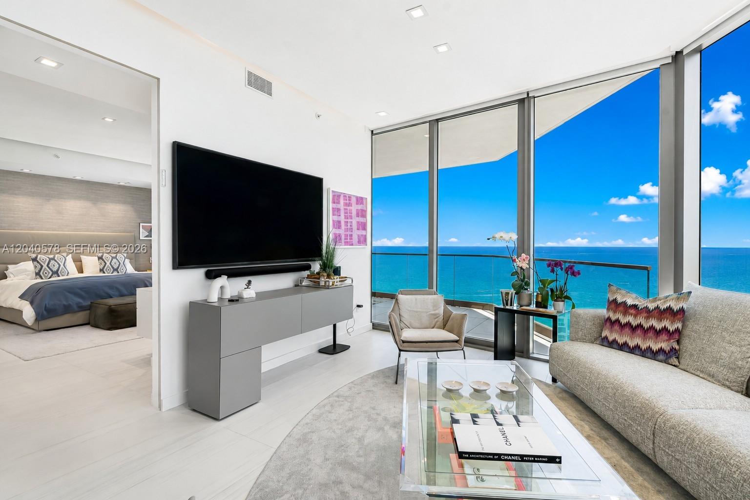 A modern living room features a wall-mounted television, gray console, and floor-to-ceiling ocean views — 18975 Collins Ave # 2004, Sunny Isles Beach FL 33160, SUNNY ISLES BEACH, FL