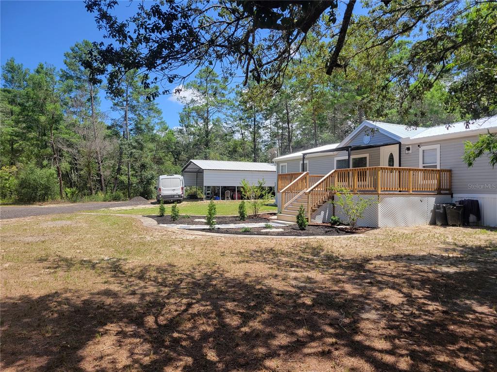 A light gray manufactured home with a wooden deck stands near a metal carport — 7597 N BRUTUS AVENUE, DUNNELLON, FL
