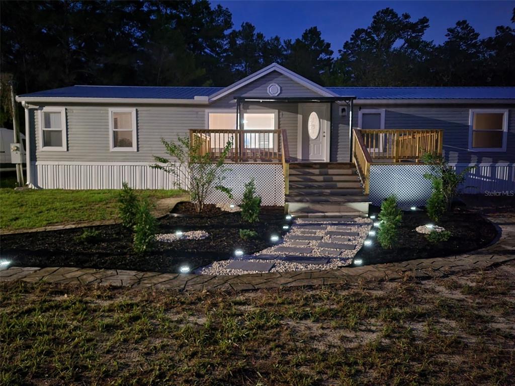 A gray manufactured home features a wooden deck, stone walkway, and landscape lighting at dusk — 7597 N BRUTUS AVENUE, DUNNELLON, FL