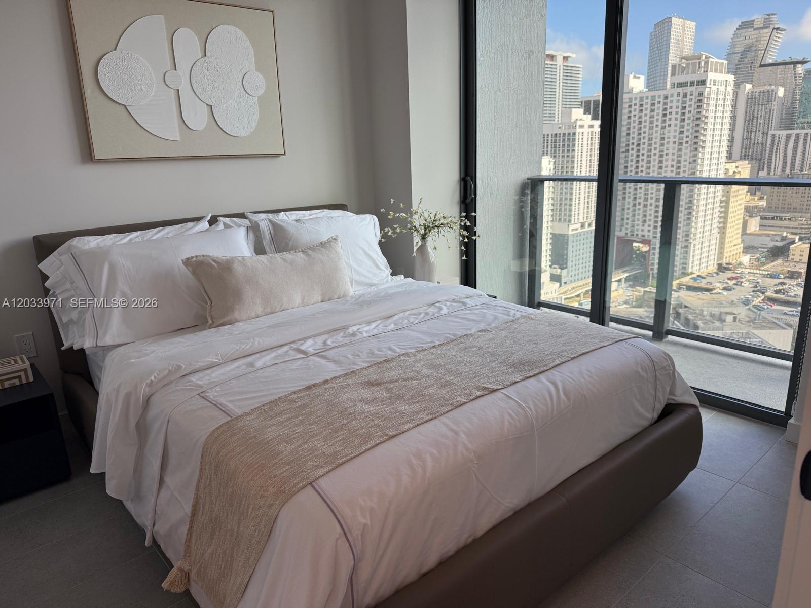 A bedroom features a bed with white linens, a textured wall art piece, and city views — 121 NE 5th St # 3002, Miami FL 33132, MIAMI, FL