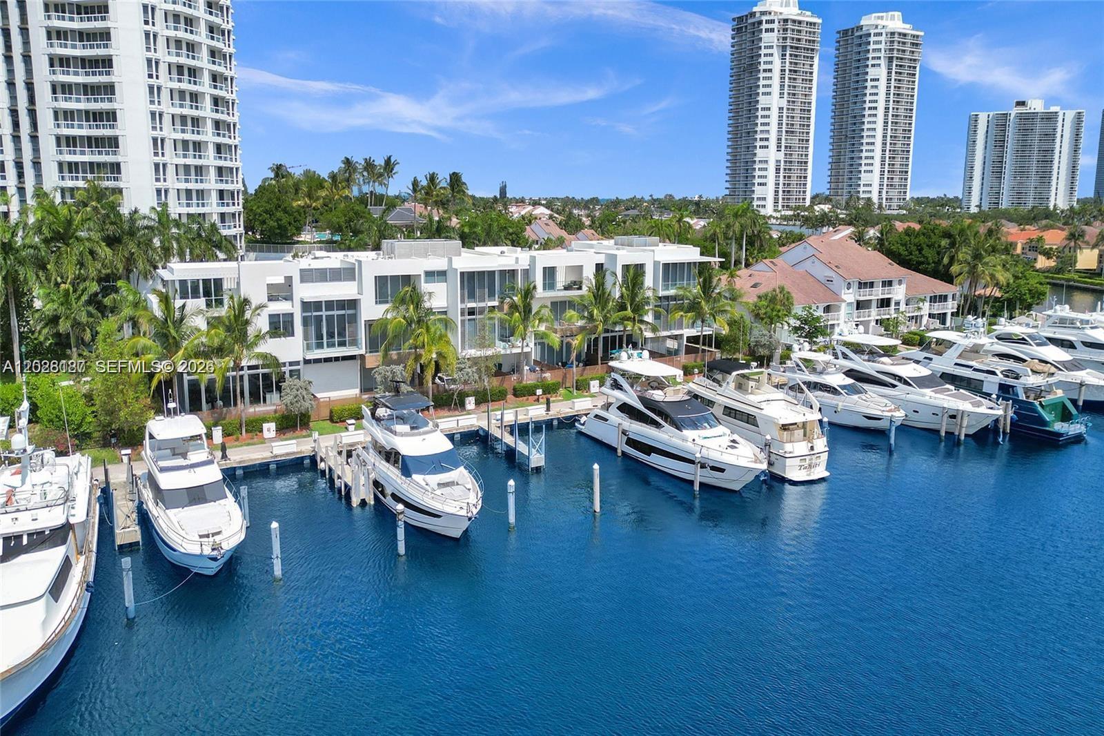 An aerial view shows a modern white building with a marina and several docked yachts — AVENTURA, FL