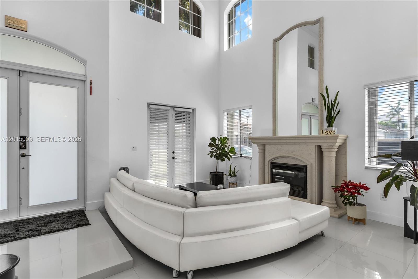 A white living room features a curved sofa, a fireplace with a mirror, and high ceilings — 15395 SW 23rd St, Miami FL 33185, MIAMI, FL