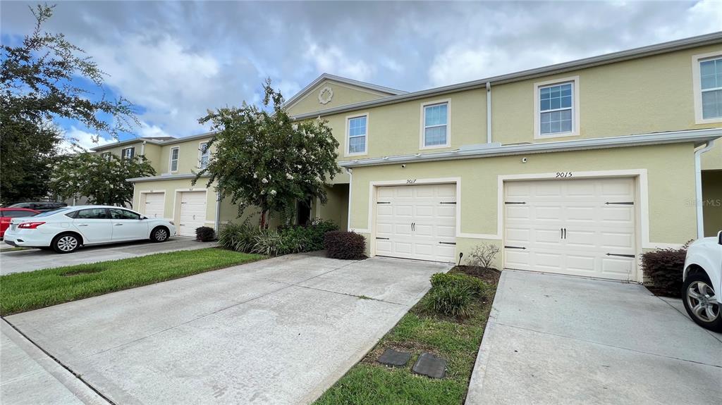 A two-story townhouse exterior features light yellow siding, white garage doors, and a concrete driveway — 9017 ALBA LANE, KISSIMMEE, FL