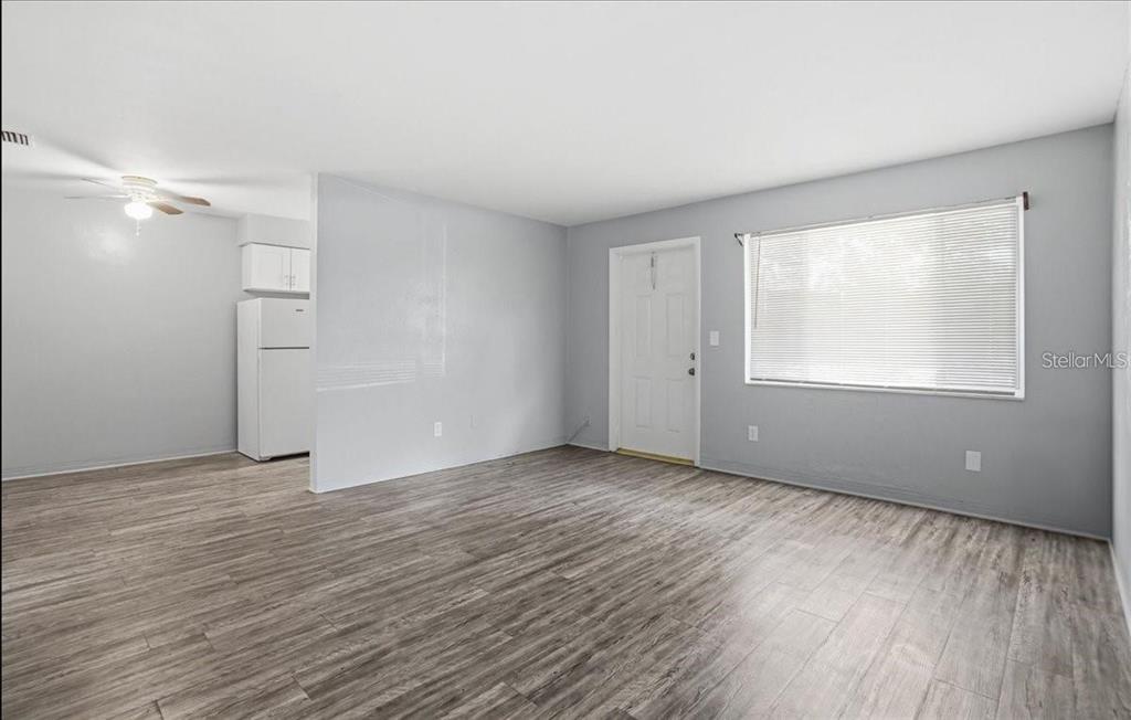 An empty living room with light gray walls, wood-look flooring, and an adjacent kitchen area — 5991 FAIRFIELD AVENUE S, GULFPORT, FL