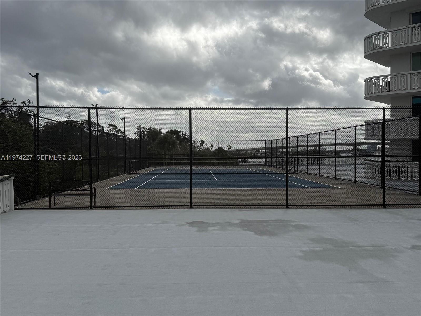 A fenced tennis court sits outdoors next to a multi-story building under a cloudy sky — 925 N Halifax # 102, Daytona FL 32118, DAYTONA, FL