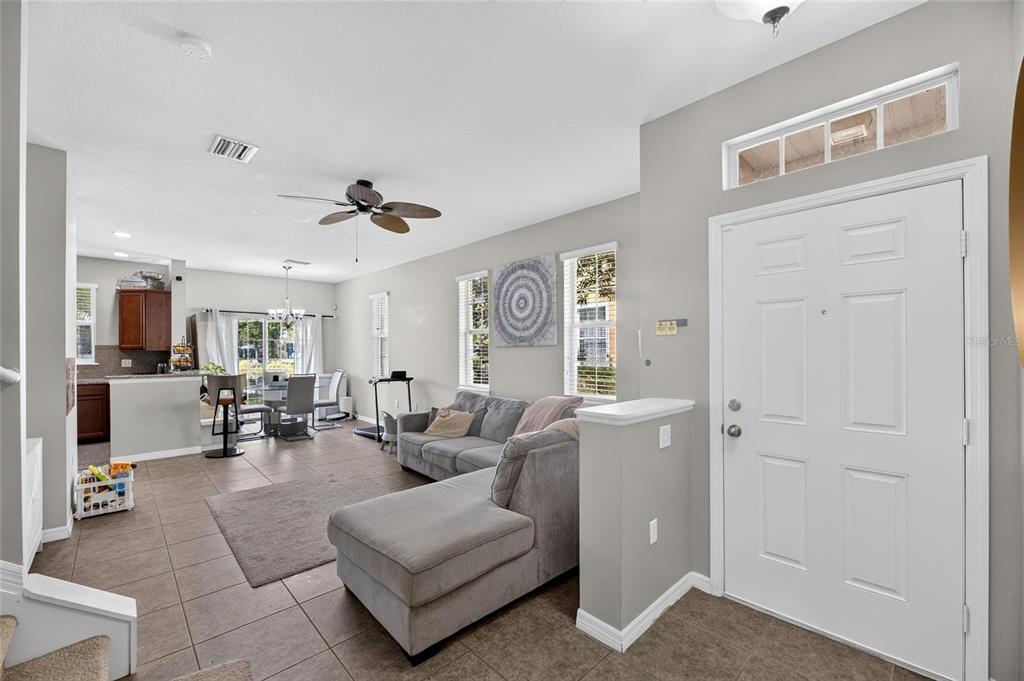 An open-concept living room with a grey sectional sofa, tile flooring, and an entryway door — 8833 MOONLIT MEADOWS LOOP, RIVERVIEW, FL