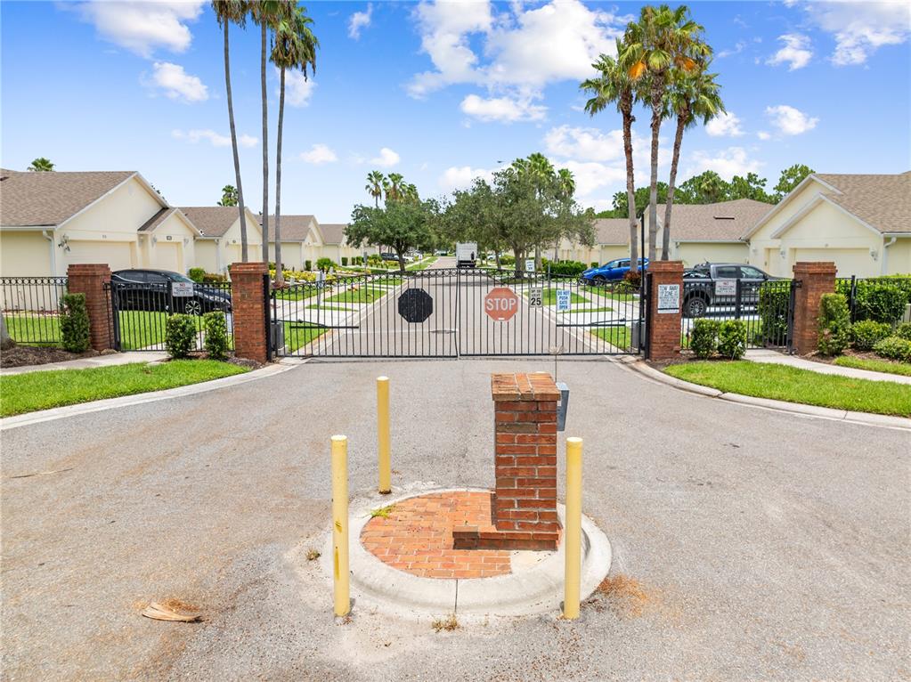 A gated entrance with brick pillars, metal fencing, and palm trees in a residential neighborhood — 153 CLUB VILLAS LANE, KISSIMMEE, FL