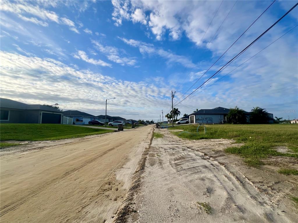 A sandy, unpaved residential street is flanked by houses and grassy lots under a blue sky — 1424 NE 9TH AVENUE, CAPE CORAL, FL
