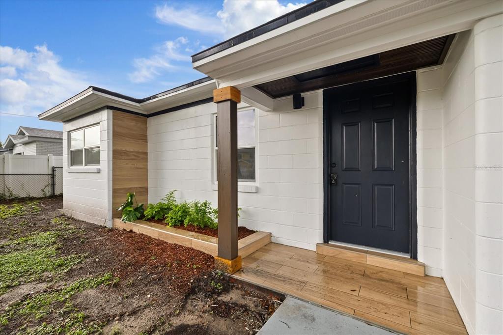 A white concrete block house exterior features a dark door, wood-look porch tiles, and landscaping — 8730 N CALDER PLACE, TAMPA, FL