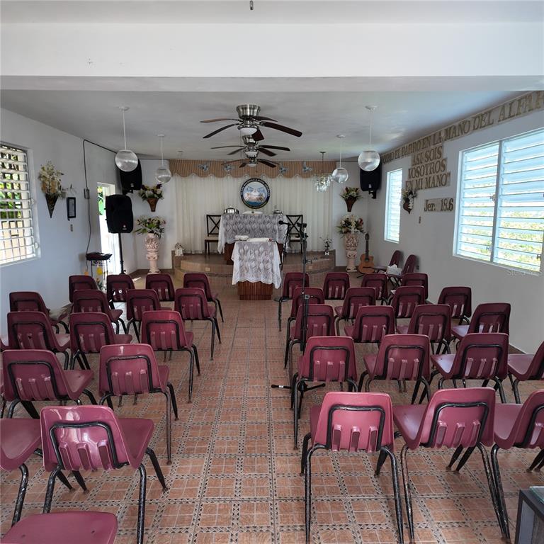 A small church interior features rows of maroon chairs facing a raised altar with curtains — Carr 157 KM 23.2 INT BO. BARROS, OROCOVIS, PR