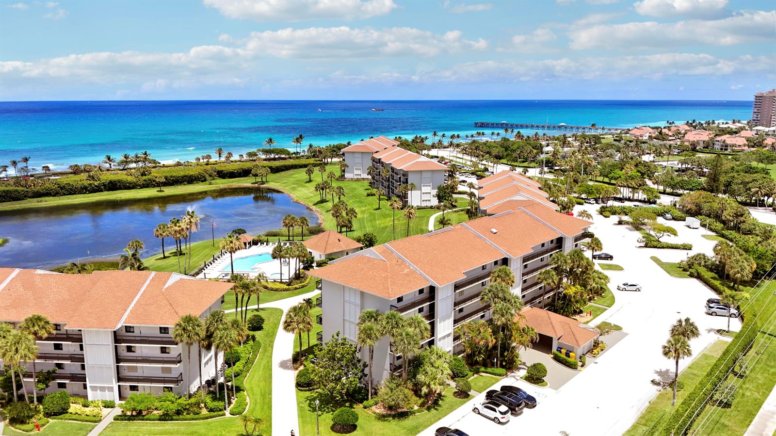 An aerial view shows multi-story apartment buildings with brown roofs near a lake and ocean — 401 S Seas Drive # 203, Jupiter FL 33477, JUPITER, FL