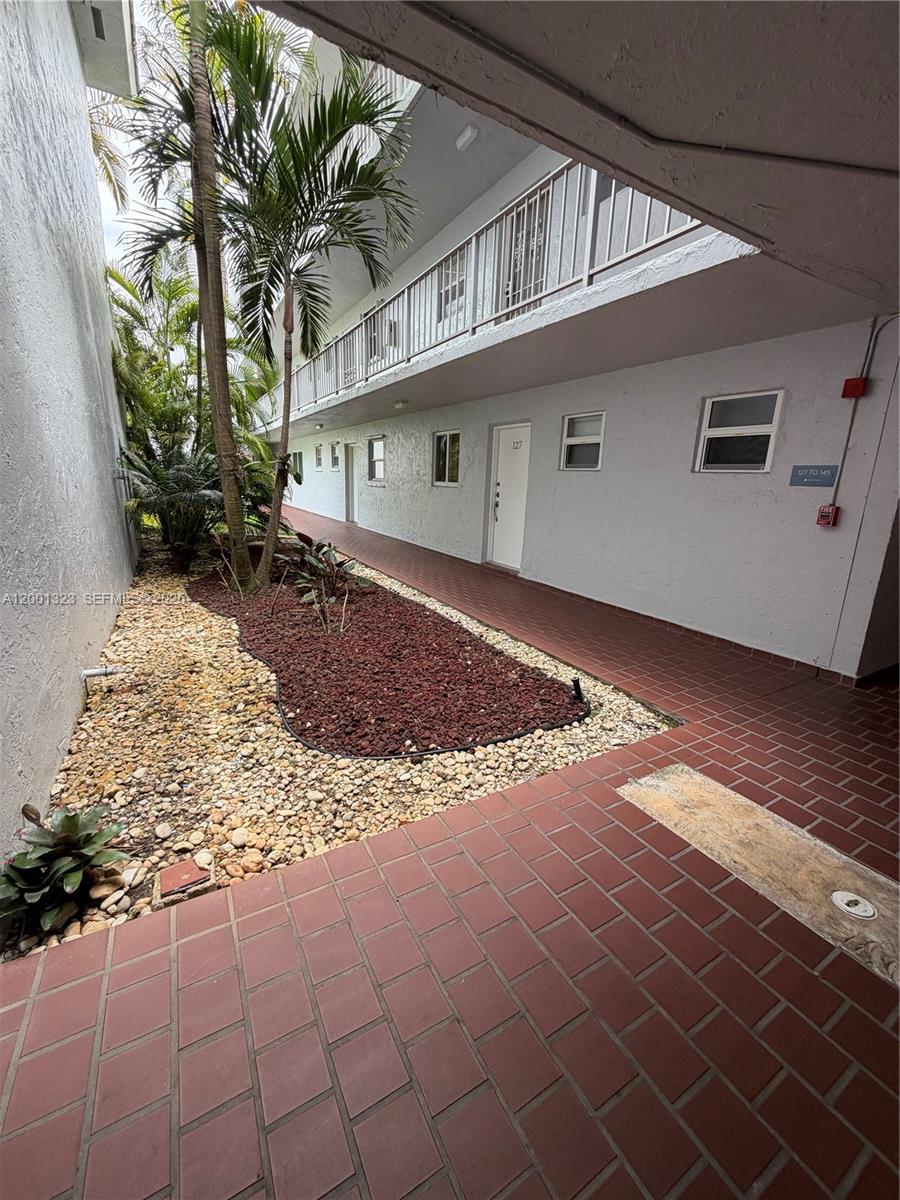 An outdoor walkway with red brick pavers leads past a garden bed toward apartment doors — 100 Edgewater Dr # 127, Coral Gables FL 33133, CORAL GABLES, FL