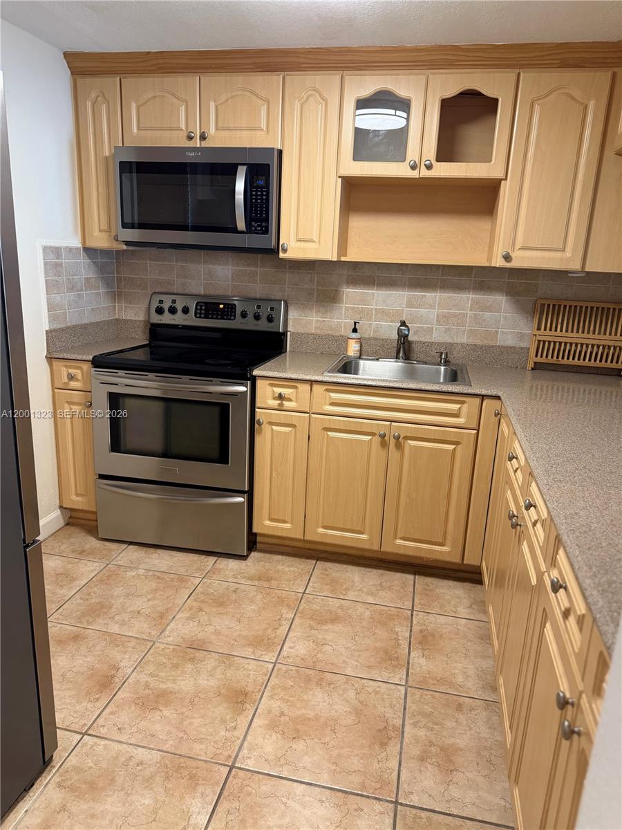 A kitchen features light wood cabinets, stainless steel appliances, a tiled backsplash, and beige flooring — 100 Edgewater Dr # 127, Coral Gables FL 33133, CORAL GABLES, FL