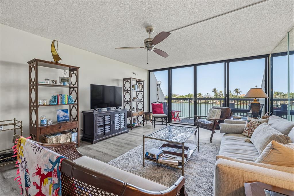 A living room features a sofa, television, shelving units, and a balcony overlooking the water — 9495 BLIND PASS ROAD UNIT 206, ST PETE BEACH, FL
