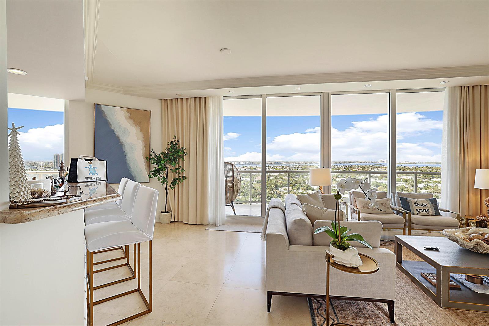A bright living room features a kitchen bar, neutral furniture, and floor-to-ceiling windows with views — 2700 N Ocean Drive # 1006b, Riviera Beach FL 33404, RIVIERA BEACH, FL